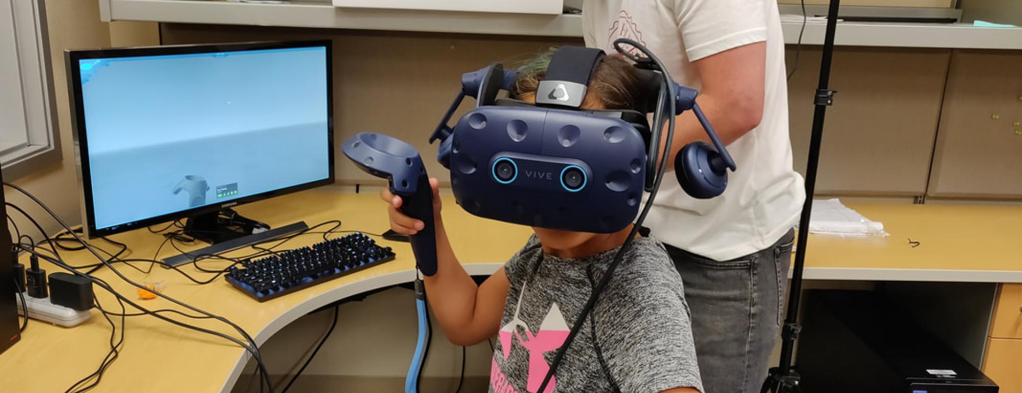 researcher with child in virtual reality goggles with joystick next to a computer