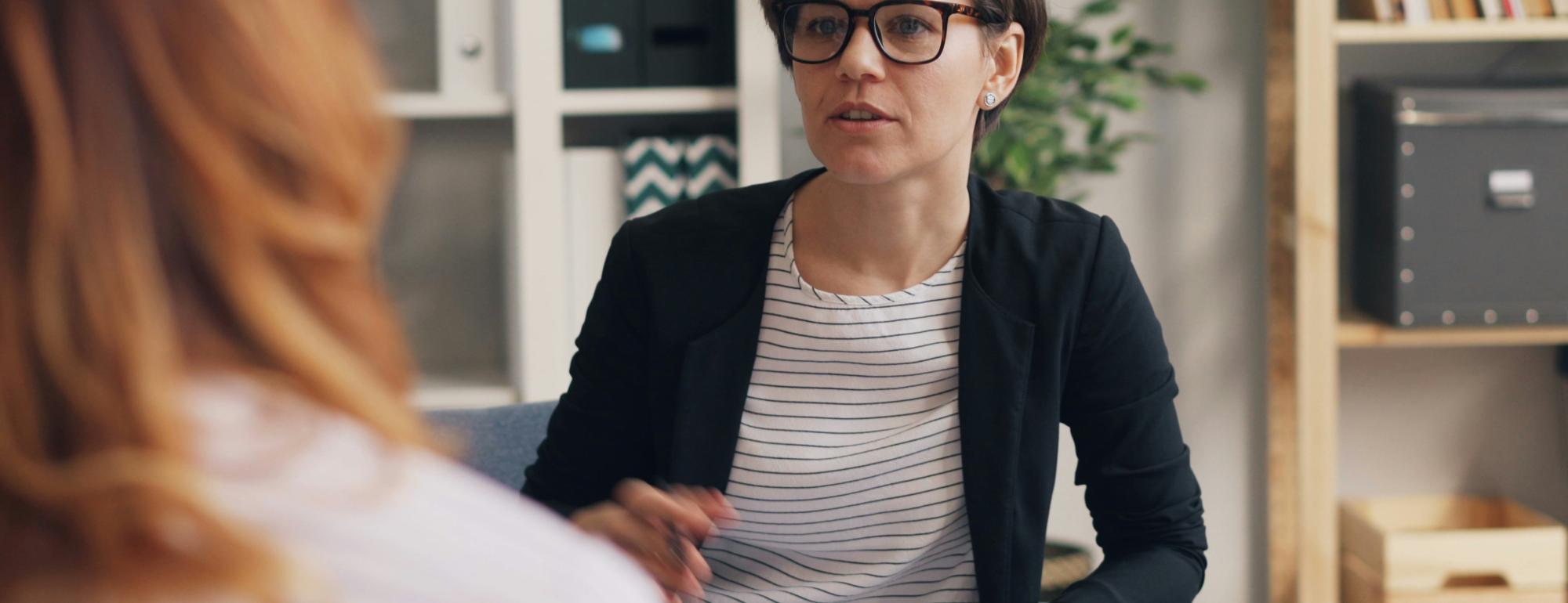 A woman in glasses talks attentively to another person in a cozy, modern office setting.