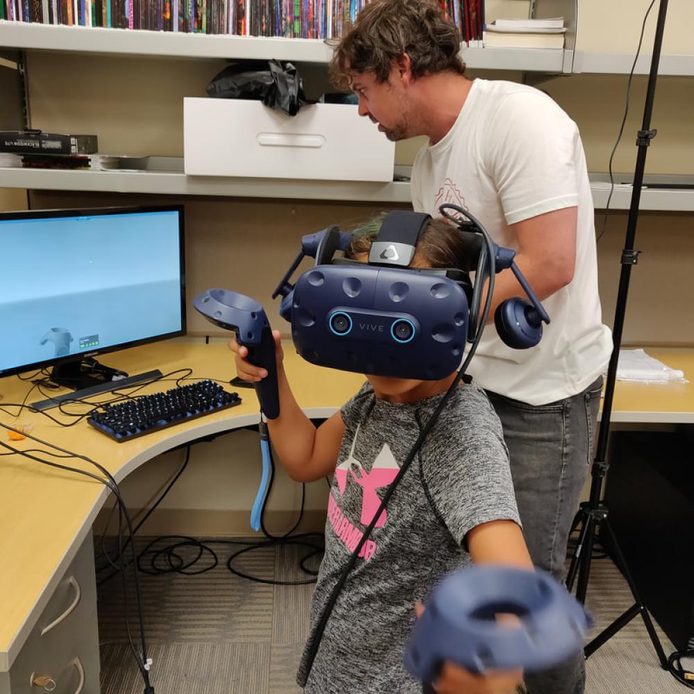 researcher with child in virtual reality goggles with joystick next to a computer