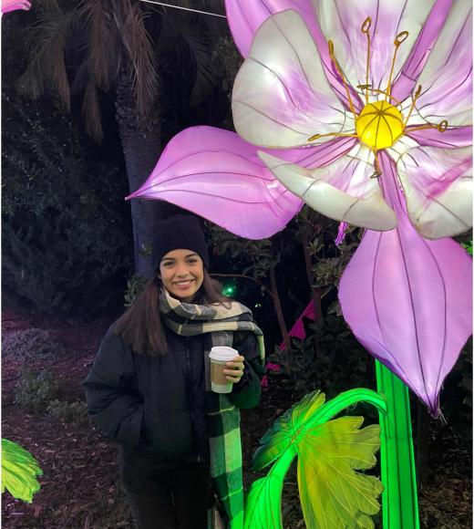 Smiling woman in winter coat holding a drink beside giant illuminated pink-and-white flower at night